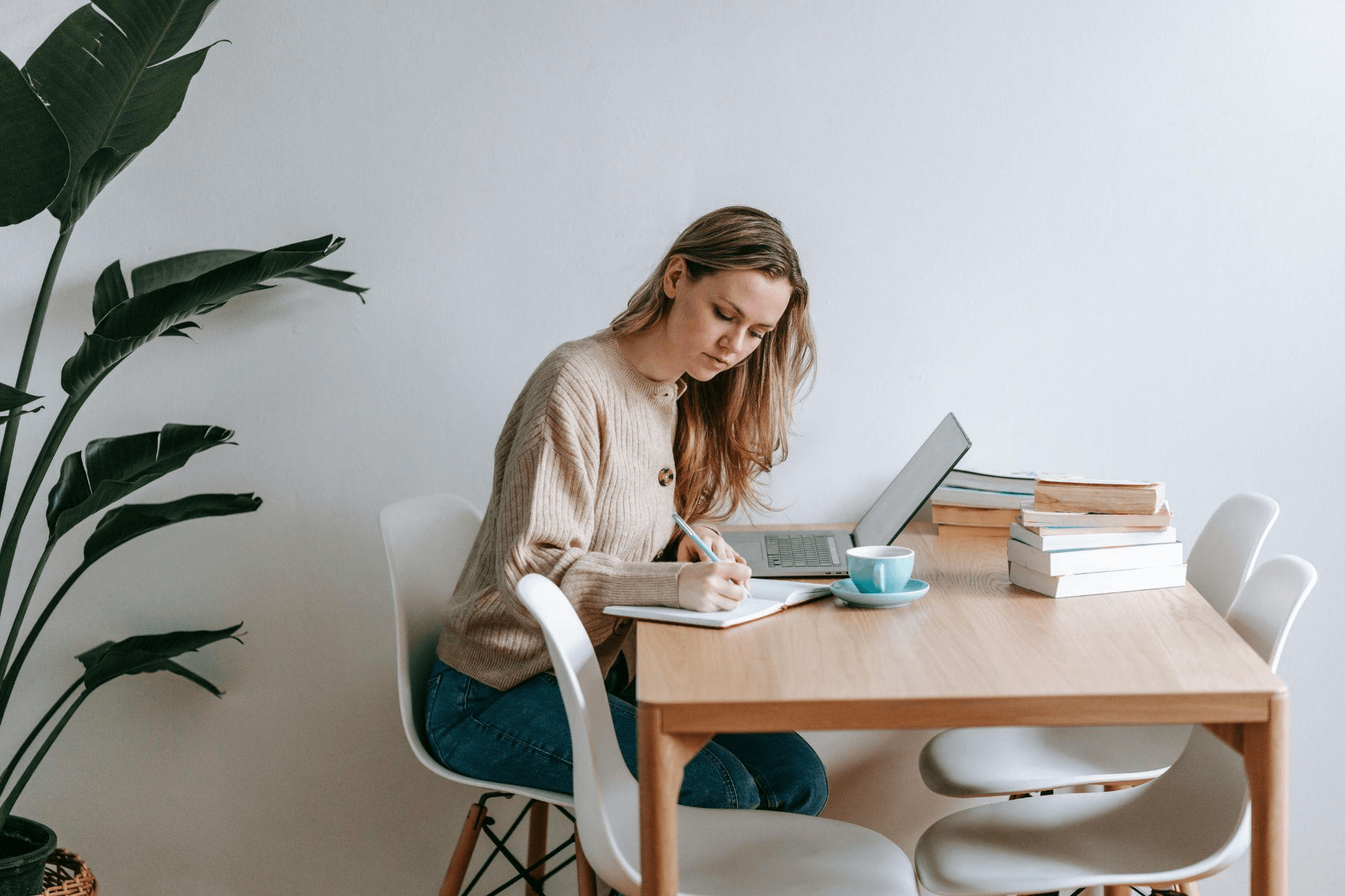 Focused woman writing in a notebook at a table with a laptop, representing a book ghostwriting service crafting a manuscript.