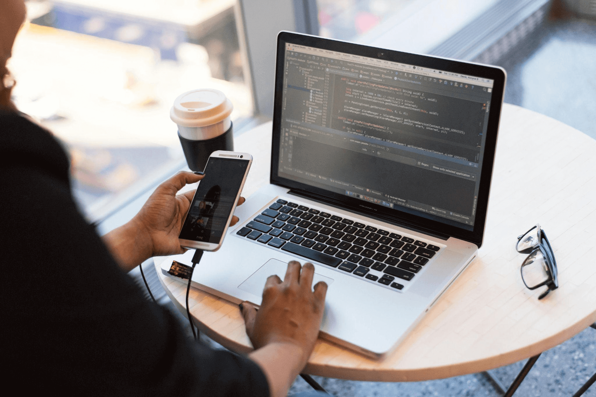 A developer holding a smartphone while working on a laptop, analyzing app performance metrics displayed on the screen. The image suggests multitasking between mobile and web platforms, emphasizing the importance of optimizing app loading times.
