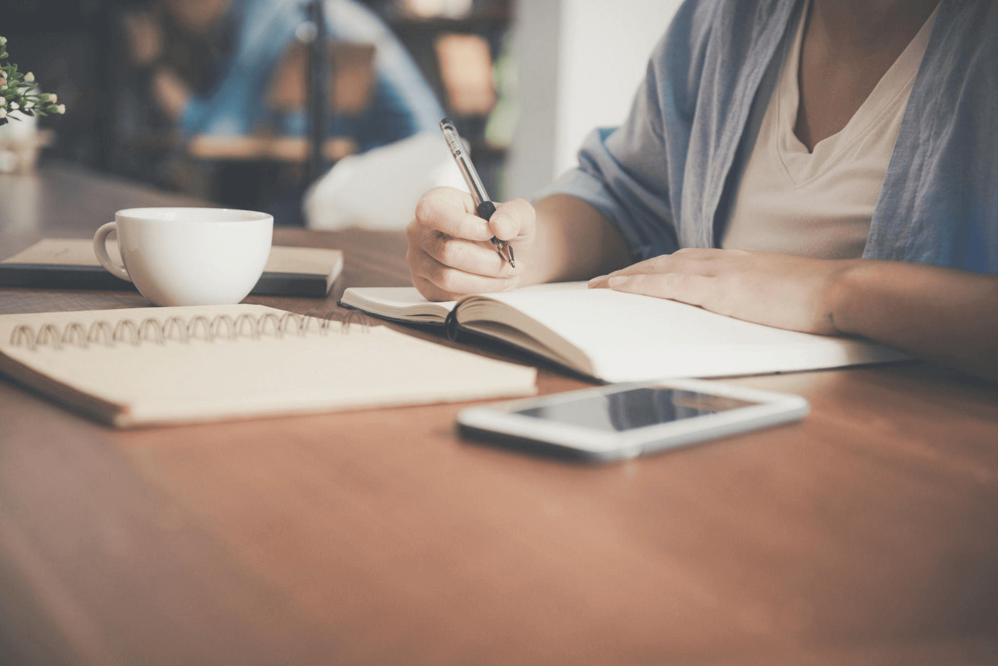 Woman writing in a notebook next to a teacup and tablet, representing the thoughtful process of a professional biography writing service.