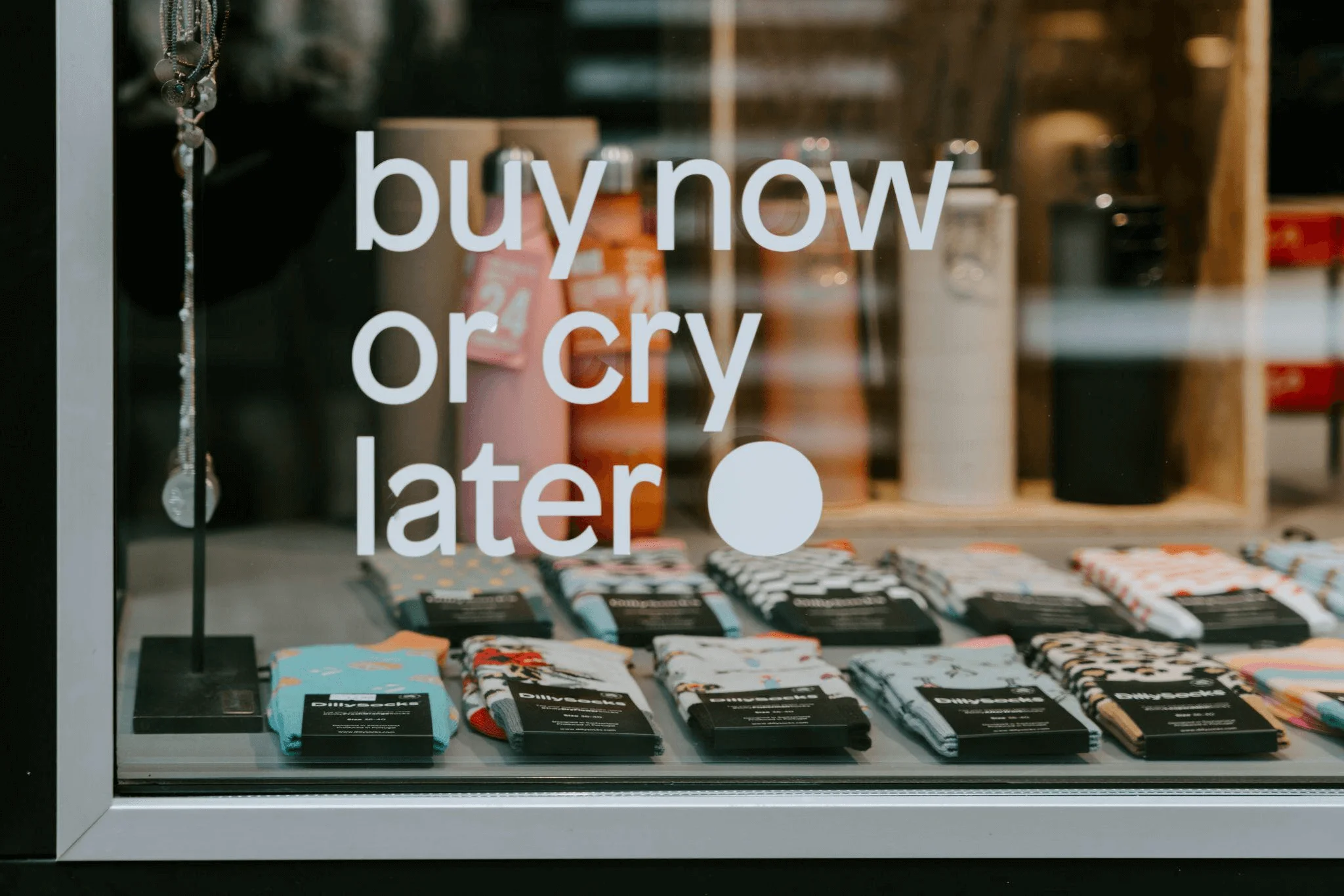 Image of a shop window that reads “buy now or cry later,” with colorful socks displayed behind the glass.